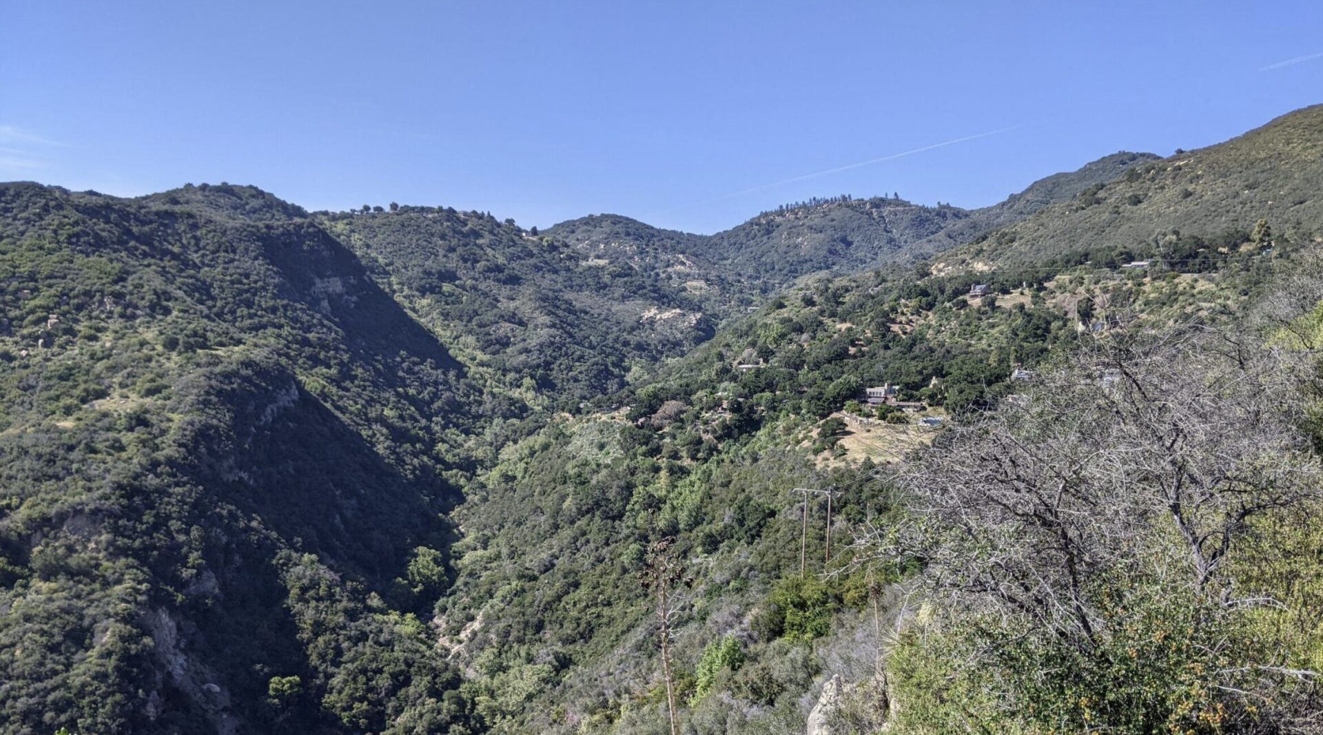 A scenic view of a hilly landscape with lush green vegetation and scattered houses nestled on the slopes, under a clear blue sky. The terrain is rugged with deep valleys and elevated peaks in the distance. Area of wildfire resistance